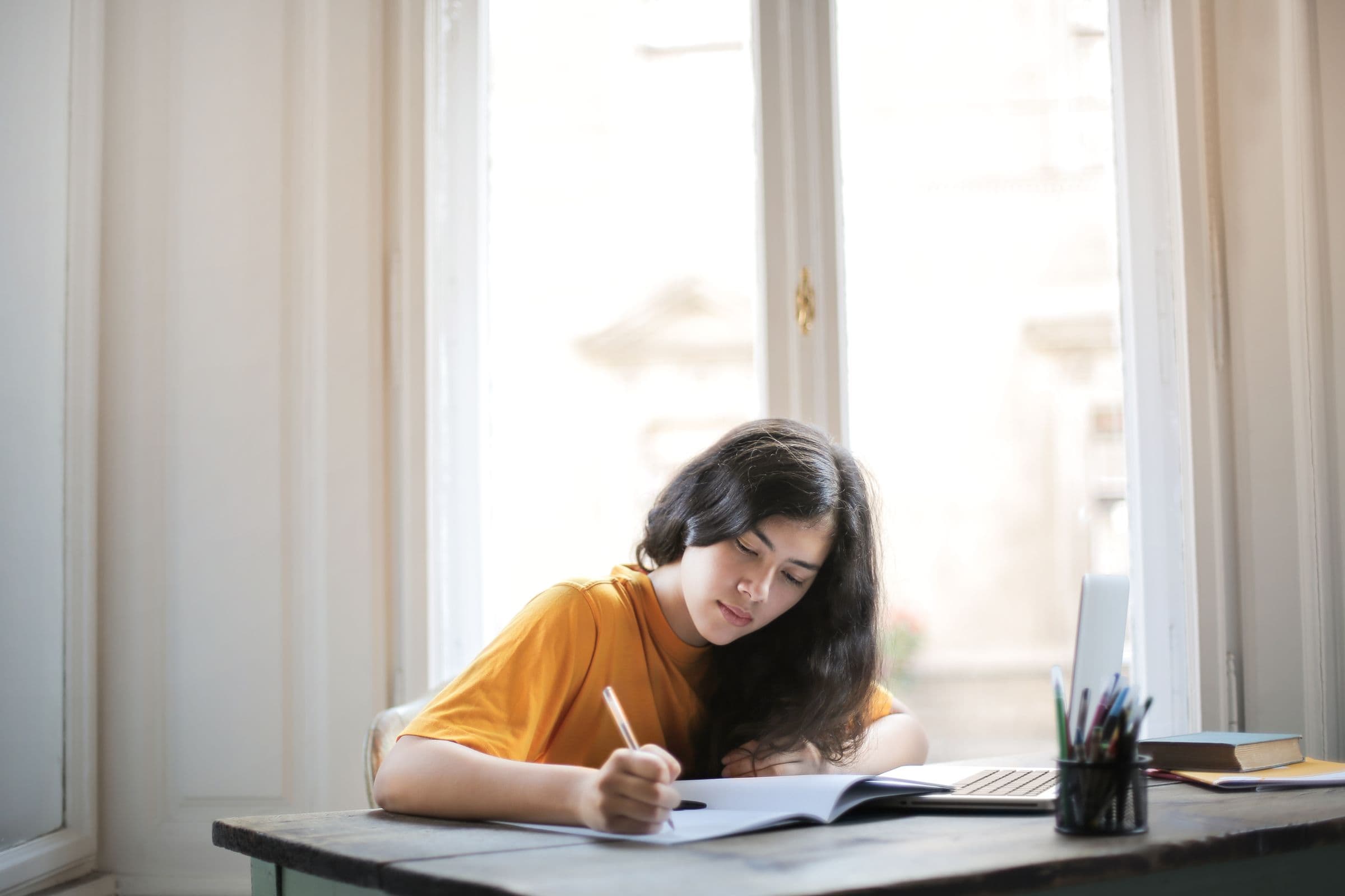 Student writing in a notebook at a sunlit desk.